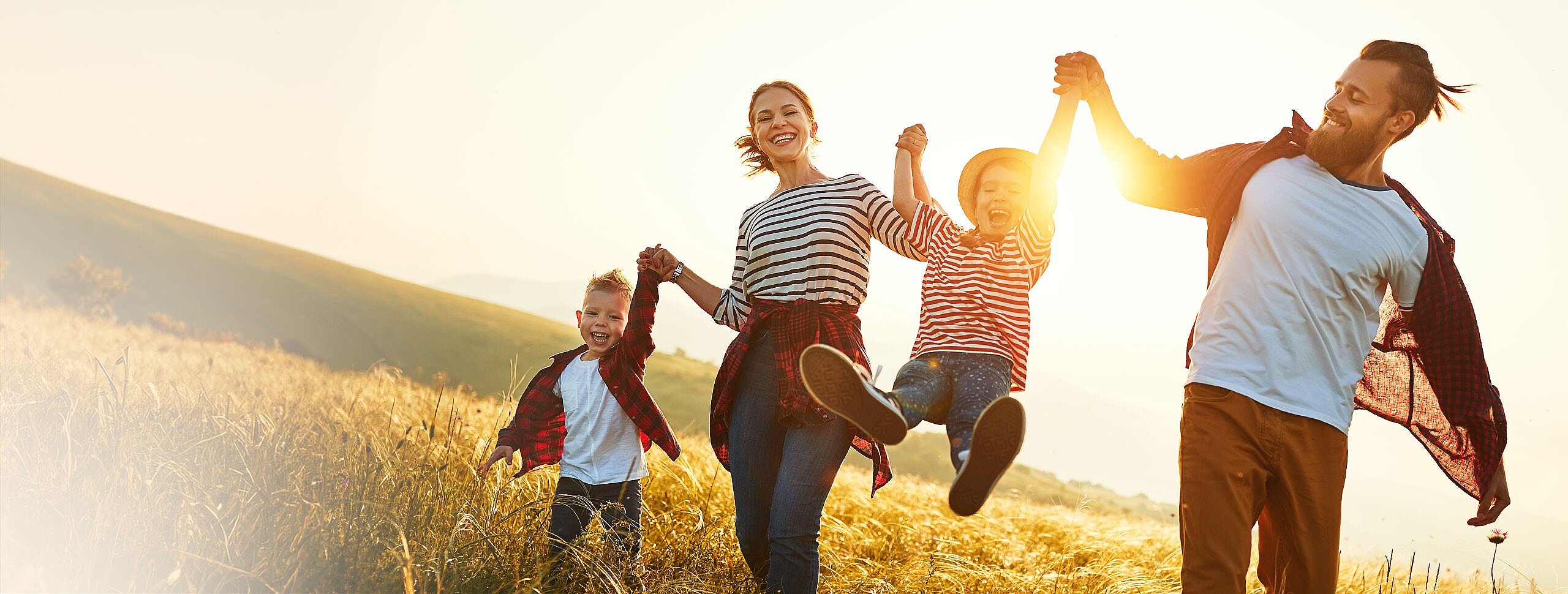 Junge Familie mit 2 Kindern rennt Hand in Hand über ein Feld und lacht. Eines der Kinde wir an den Händen hochgehoben.